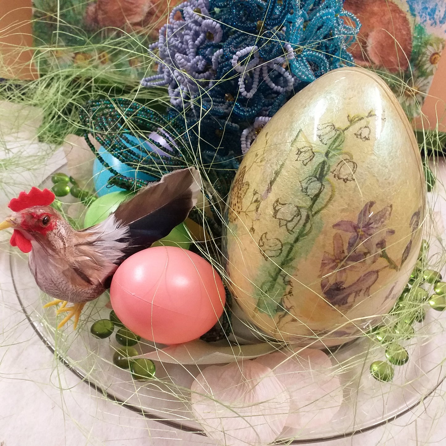 dall'alto piatto di vetro idea regalo centrotavola pasquale ortensia fiori di perline azzurri blu ovetti di plastica gallo piume decorazioni addobbi petali sassolini vetro uovo grande mughetti violette sfondo vetrina runner coniglietti